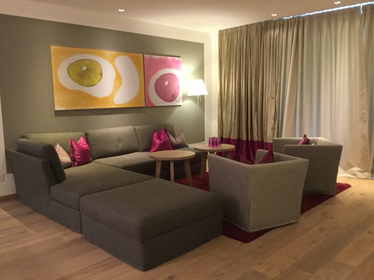 Dolomitengolf Suites Golfhotel, Blick in eine Juniorsuite mit grauem Sofa und pinken Kissen.